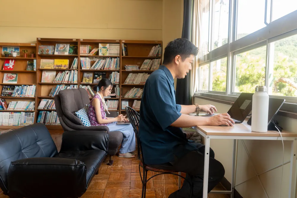 本棚にかこまれた元図書室で、窓ぎわの机に向かう人とソファで本を読む人